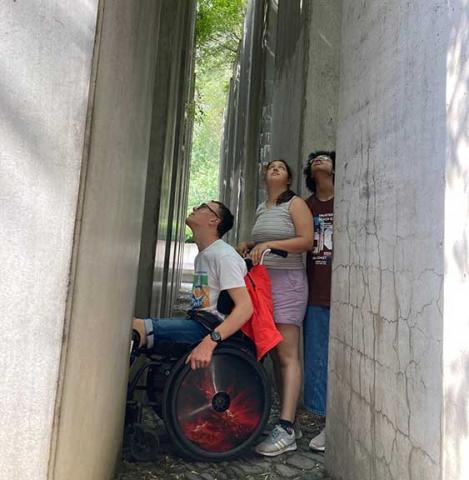 The picture shows three teenagers, one of them using a wheel chair. They are squeezed in between blocks of concrete and looking upwards.