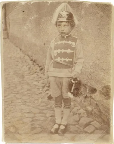 Photography of Walter Frankenstein as a child, costumed on cobblestones standing in front of a house wall.