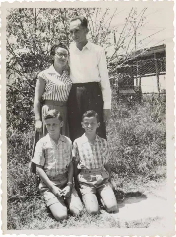 The photo shows a man, a woman and two boys outdoors. The children kneel on the floor. On the left behind them is the woman, on the right next to her is the man. In the background there is a garden and a building.