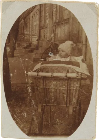 The portrait-format, oval vignetted photograph shows an infant (Walter Frankenstein) in a pram made of braided basket.
