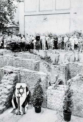 Black and white photograph: Amidst the remains of a wall, a man fits a brick into a wall. In front of him on the left is a lectern decorated with an Israeli flag and a microphone. Spectators stand in the background.