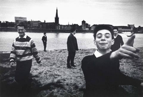 Black and white photograph: Six young people stand on the banks of a wide river. They are not standing together, but spread out across the entire picture. Some are looking towards the viewer, others across to the city that stretches along the opposite side of the river. The group makes an exuberant impression.