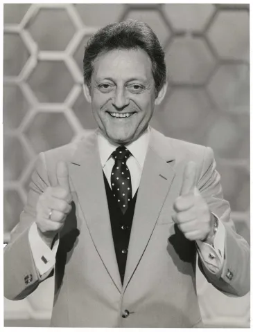 The black and white photo shows a half-portrait of a man in suit and tie with both thumbs up and looking friendly into the camera, with honeycomb pattern in the background.