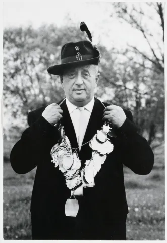 Black and white photograph: A gentleman in a hunting hat and suit presents a necklace with numerous pendants.