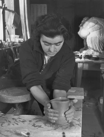 Black and white photo of a woman at the pottery wheel shaping a cup with her clay covered hands