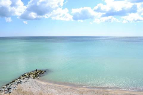 Leichte Wolken über türkis-blauem Meer, am vorderen Bildrand ein Stück Strand.