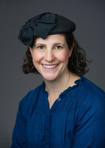 Portrait of a young woman smiling into the camera with shoulder-length dark curls, black hat and blue blouse.