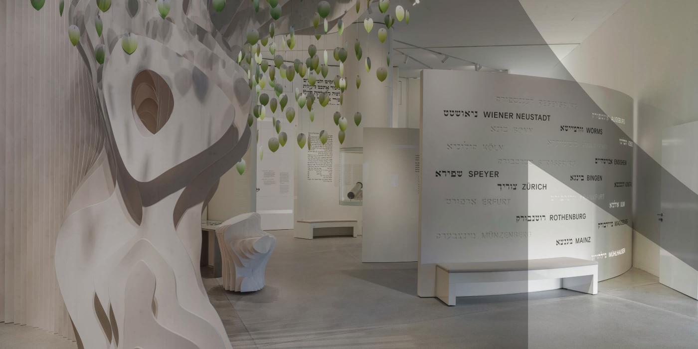 Ausstellungsraum in weißem Design, im Vordergrund ein aus weißen Elementen nachempfundener Baum. Das Bild ist mit einem grauen Schleier der das JMB-Logo andeutet.