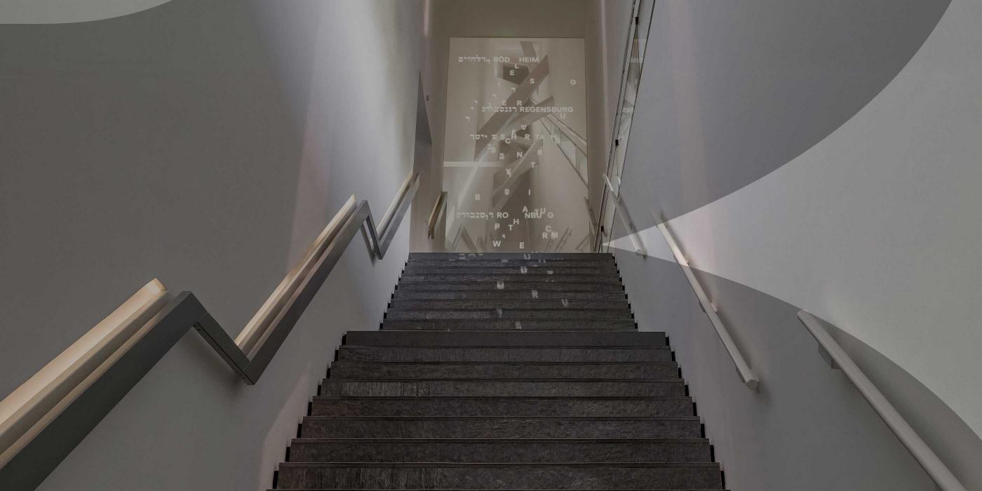 Staircase in a new building, at the end of which is a projection with white words in Hebrew and German. The image is with a gray veil that suggests the JMB logo.