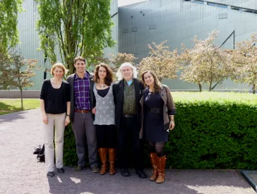 A group of adults is standing next to a hedge in front of a modern building. Trees can be seen in the background.