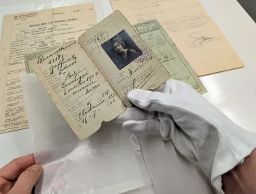 A hand with a cotton glove holds a historical identity card up to the camera. Other historical documents can be seen in the background.