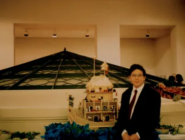 A boy in a dark suit and tie stands next to a toy ark made of wood. A rotunda is visible in the background.
