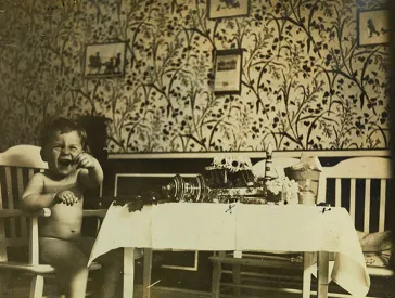 Boy sits naked and a little upset at the table; the living room is wallpapered with a floral wallpaper.