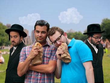 Two men in short-sleeved shirts are standing in the park, eating baked goods out of paper bags and looking at the camera; next to them are two men dressed in Jewish Orthodox clothing.