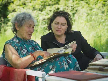 Two women sit on a garden bench and leaf through a photo album together.