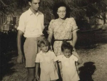 The photo shows a man, a woman and two children in front of them, all standing outside. The two boys and the man are wearing shorts, the woman a short-sleeved dress with a floral pattern.