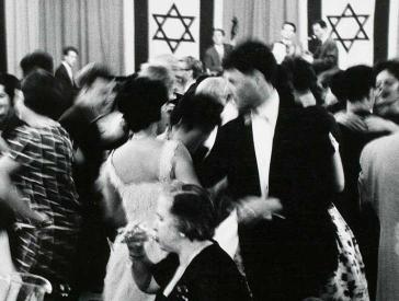 Black and white photograph: people dressed in festive outfits dancing; in the background is a stage decorated with two Israeli flags and a band playing.