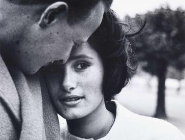 Black and white photograph: Portrait of a young couple: She leans her face against his shoulder and looks into the camera, his face is turned towards her and can be seen in profile from diagonally behind, his head is leaning against hers.