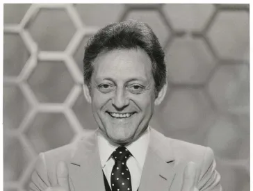 The black and white photo shows a half-portrait of a man in suit and tie with both thumbs up and looking friendly into the camera, with honeycomb pattern in the background.