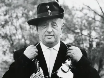Black and white photograph: A gentleman in a hunting hat and suit presents a necklace with numerous pendants.