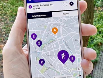 Eine Hand hält ein Smartphone, auf dem eine Karte mit bunten Ortsmarkern geöffnet ist.