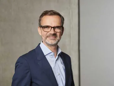 A man with brown hair and glasses stands in front of a concrete background.