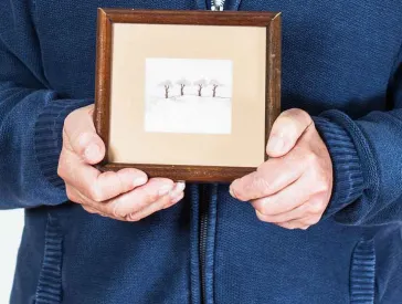 Elderly man holding a drawn picture with four trees in a frame in his hands.