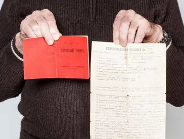 A bearded, gray-haired man holds up two documents.