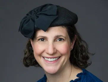 Portrait of a young woman smiling into the camera with shoulder-length dark curls, black hat and blue blouse.