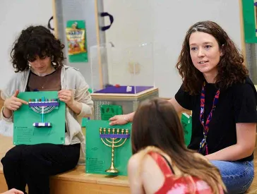 Two woman hold green squares to which Hanukkah Menorahs are attached.