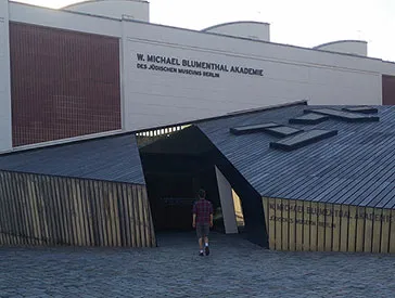 A person in shorts walks towards the entrance of the Academy of the Jewish Museum. The person is seen from behind.
