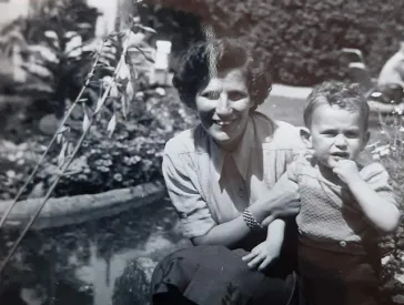 Black and white photo of a toddler in leather pants, next to him kneels a woman in skirt and blouse, in the background a house and many plants.