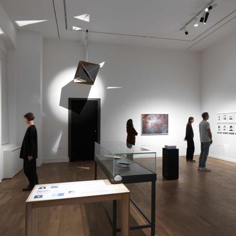 View of an exhibition room with visitors looking at paintings and photos on the walls.