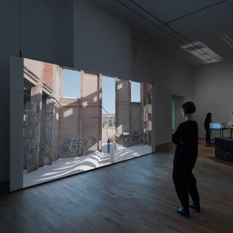 Visitors look at a large screen on which a long-haired person wearing jeans and a bare upper body is walking through the snow in a dilapidated brick building.