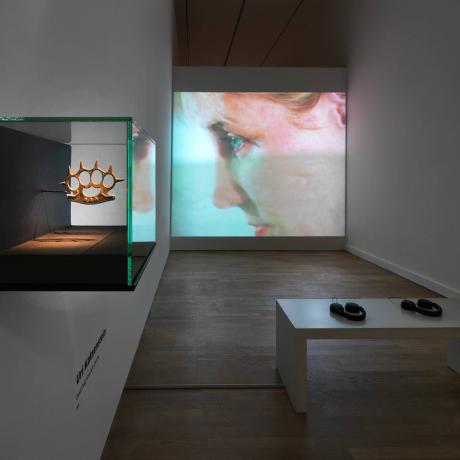 View into an exhibition room, in the foreground a display case in which a knuckleduster is exhibited, in the background a screen on which a close-up of a face can be seen, in the middle of the room a bench with two headphones.