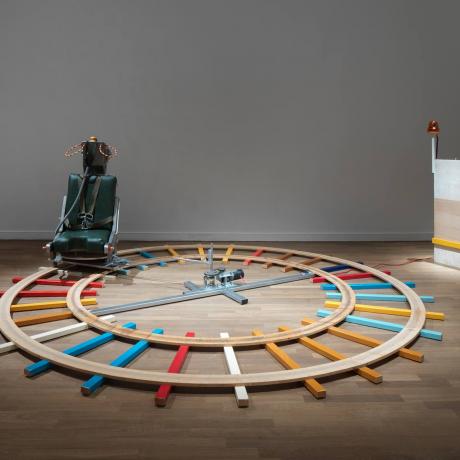 Exhibition room with colorful wooden rails running in a circle on the floor, with an ejection seat on top and on the right a wooden desk with an orange light wired to the seat.