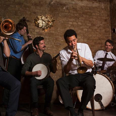 Six male members of a jazz combo play their insturments in front of a brick wall.