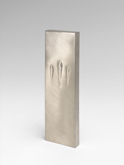 The image shows the art work Touching Mezuzah by Sari Srulovitch. The object ha s a rectangular shape and is made of silver. 