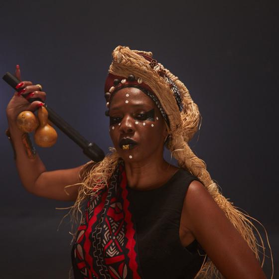 Female and Caribbean read person with red and black sash, straw headdress and stick with calabash in hand against a dark background.