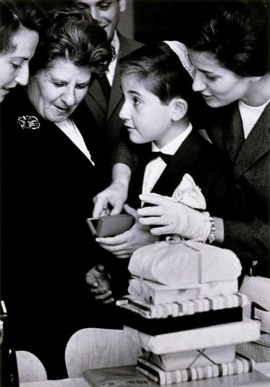 Black and white photograph: In the center of the photo is a boy in a suit and with a white kippah in profile, surrounded by three women and a man. A pile of presents can be seen in front of the boy.