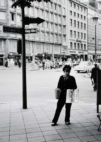 Schwarzweiß-Fotografie: Ein Zeitungsverkäufer, der eine Schirmmütze trägt, steht mit Zeitungen unter dem Arm an einer Kreuzung.