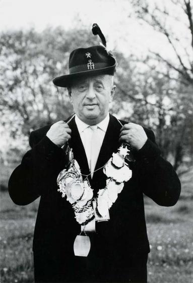 Black and white photograph: A gentleman in a hunting hat and suit presents a necklace with numerous pendants.