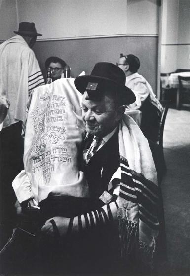 Black and white photograph: An elderly man carries a Torah scroll covered with a Torah mantle. He himself is wearing a hat as a head covering, a tallit and a prayer strap around his arm. Behind him, three other similarly dressed men can be seen in conversation.