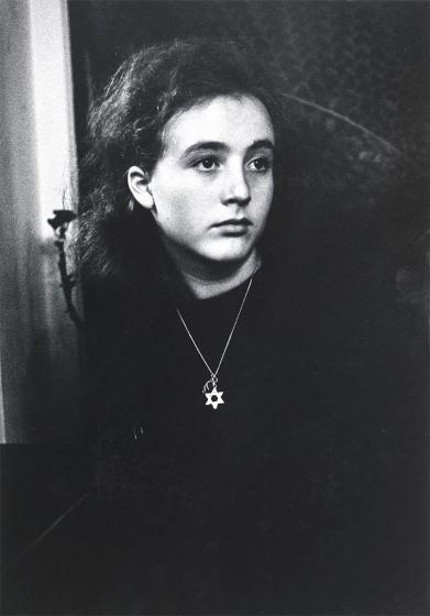 Black and white photograph: Portrait of a girl in half-profile facing right. She is wearing dark clothing and dark wavy long hair. She wears a necklace with a Star of David pendant around her neck.