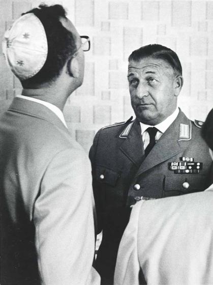 Black and white photograph: A man with a kippah can be seen in the foreground of the picture from diagonally behind, his head and gaze directed upwards. He is standing in front of a middle-aged man in uniform, who can be seen on the right of the picture in three-quarter profile. His gaze is also directed upwards. Both are standing in front of a wall with wallpaper patterned with rectangular shapes of different sizes.