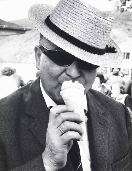 Black and white photograph: A middle-aged man with sunglasses and a sun hat is eating an ice cream. In the background, many people can be seen in a blur.