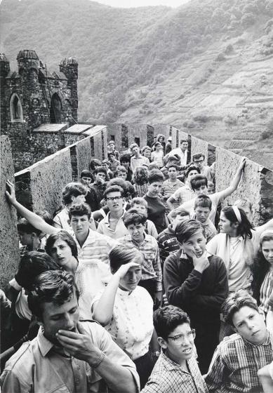 Black and white photograph: A group of young people stand on the defensive wall of a castle. The photographer is taking the picture from a slightly elevated position. In the background you can see a defense tower on the left and vineyards in a hilly landscape on the right.