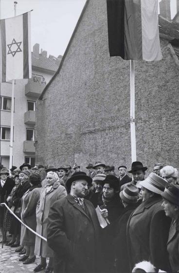 Black and white photograph: A crowd stands behind a cordon in front of a house wall. Most of the people are wearing coats and hats. An Israeli and a German flag are hoisted in the upper part of the picture. In the background is an apartment building with balconies; a man and a woman are standing on the top one and looking down.