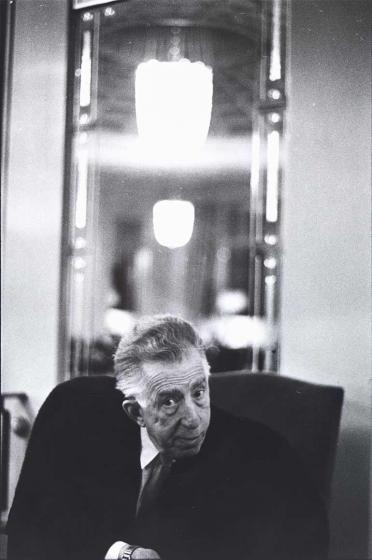 Black and white photograph: An older man with gray hair in a dark suit sits in an armchair and looks into the camera from a slight angle. A tall mirror can be seen in the blurred background.