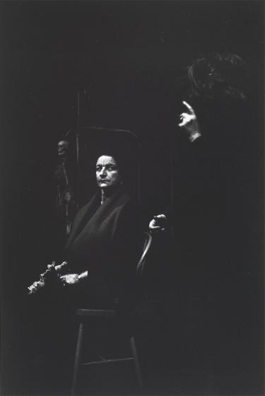 Black and white photograph: An older woman in dark clothing and with dark hair is sitting on a chair. Next to her stands another woman, she raises her index finger, the two are looking at each other. A skeleton is poorly visible in the background, possibly a dimly lit scene on a theater stage.